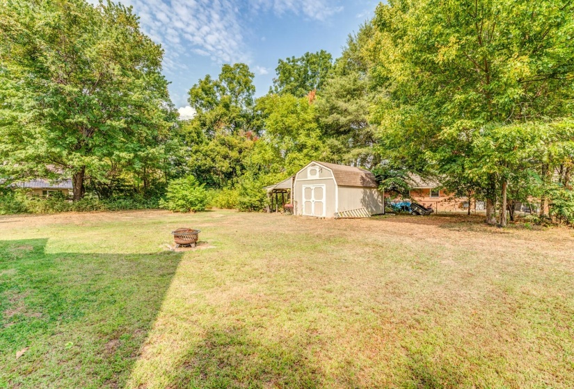 View of green lawn featuring a storage unit, a fire pit, and view of wooded area