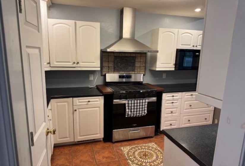 Kitchen featuring a stainless steel range with a ventilation hood, black countertops, white cabinetry with gold-toned hardware, and a tile backsplash