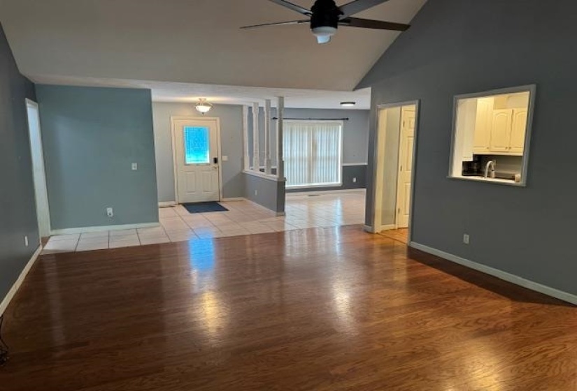 Spacious living area featuring wood-finish flooring, vaulted ceiling with a ceiling fan, and a pass-through window to the kitchen