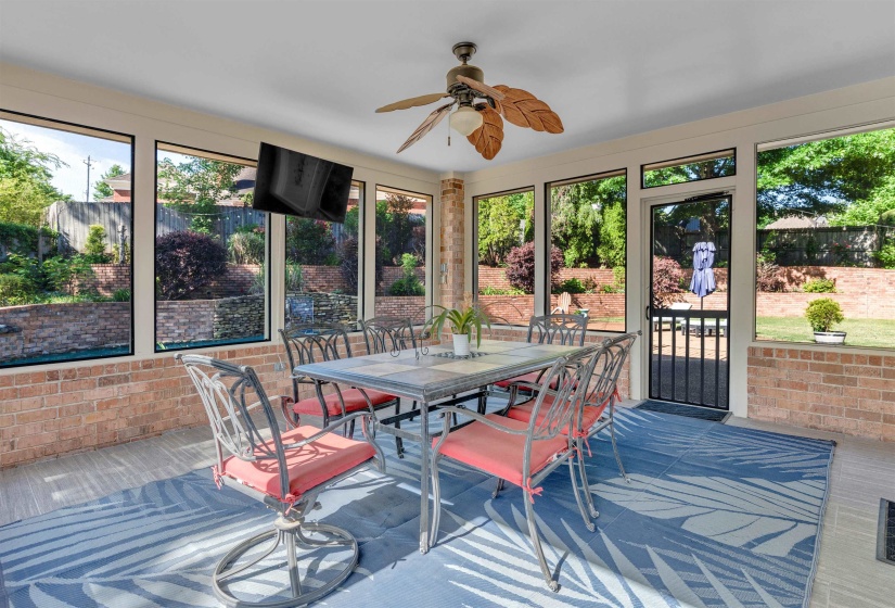 Enclosed patio featuring a ceiling fan with leaf-shaped blades, integrated wall-mounted television, and a brick half-wall surround