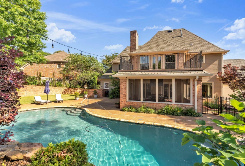 Curved swimming pool with an exposed aggregate deck, featuring a screened porch, upper balcony with iron railings, and a brick chimney