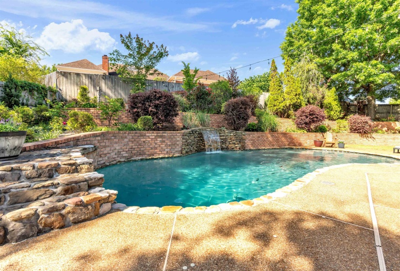 Curved swimming pool with a stone waterfall feature, surrounded by exposed aggregate concrete decking and a terraced brick retaining wall