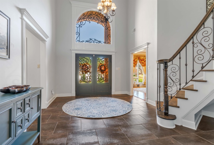 Grand foyer featuring double entry doors with decorative ironwork, an arched transom window with matching iron detailing, a wrought iron staircase with wood-finish handrail, and large format tile flooring