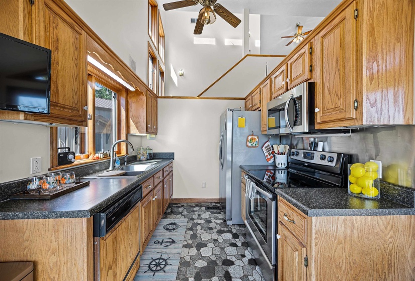 Kitchen with stainless steel appliances, dark countertops, brown cabinetry, ceiling fan, and a towering ceiling