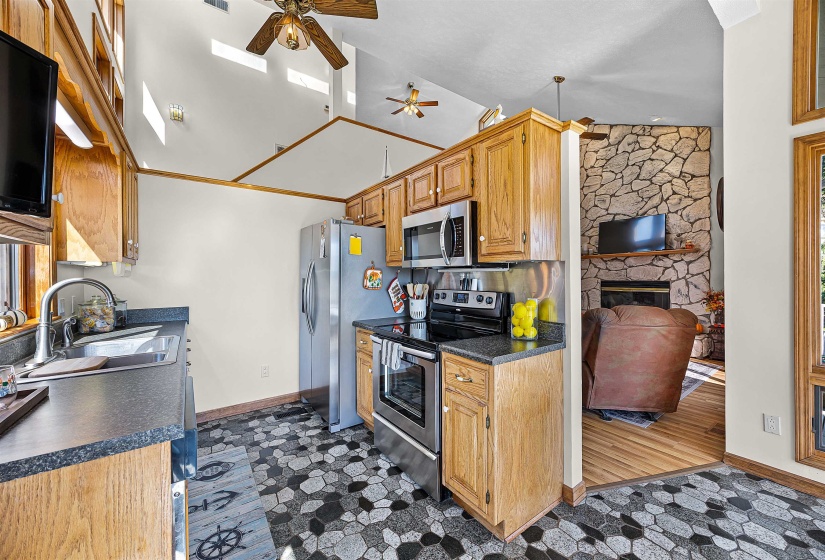 Kitchen featuring dark countertops, stainless steel appliances, ceiling fan, a fireplace, and high vaulted ceiling