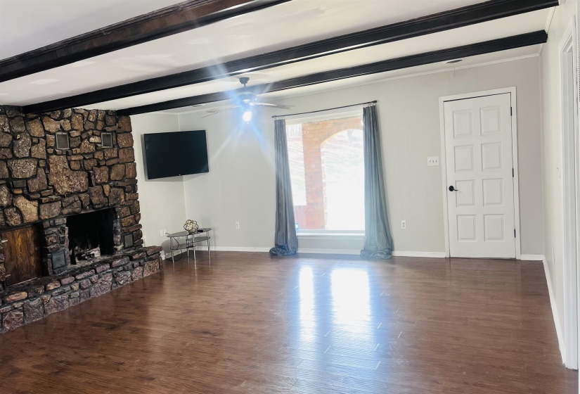 Unfurnished living room featuring dark wood-style flooring, a stone fireplace, beam ceiling, and ceiling fan