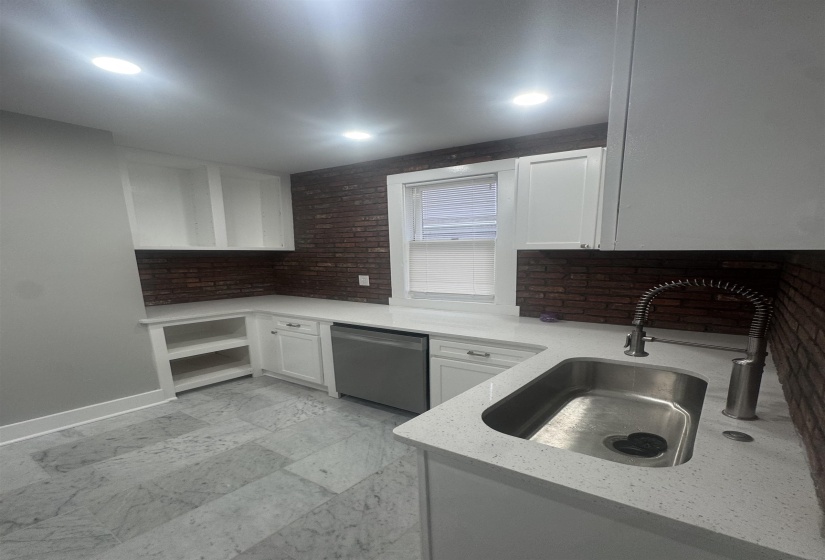Kitchen with white cabinets, dishwasher, light stone counters, open shelves, and brick wall