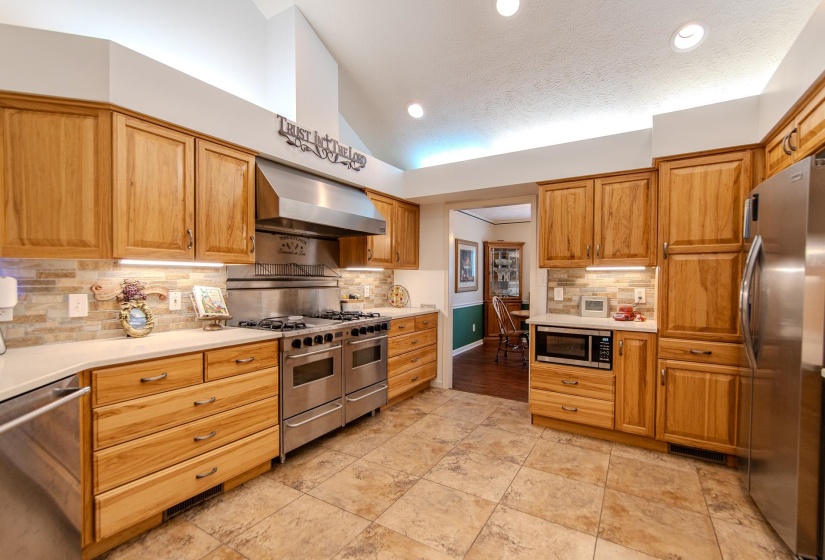 Kitchen featuring stainless steel appliances, decorative backsplash, ventilation hood, vaulted ceiling, and brown cabinetry