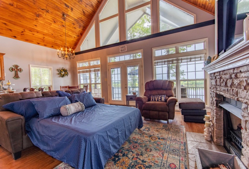Living room with high vaulted ceiling, light wood-style floors, a stone fireplace, multiple windows, and wooden ceiling