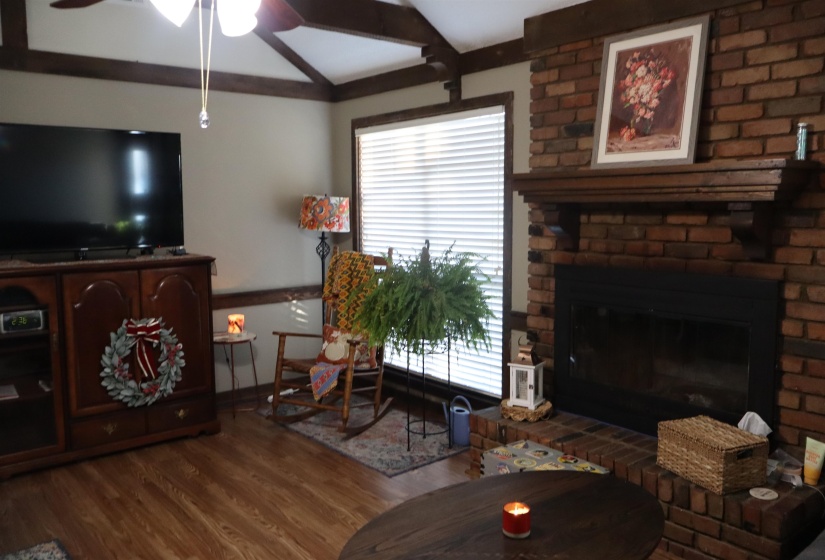 Living room with dark wood-style flooring, a brick fireplace, a ceiling fan, and beam ceiling