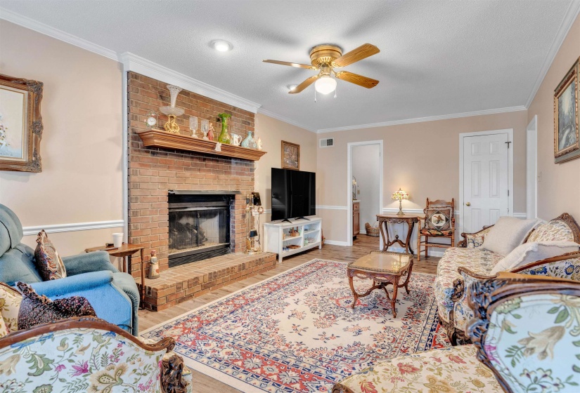 Living area with crown molding, wood finished floors, a brick fireplace, a textured ceiling, and ceiling fan