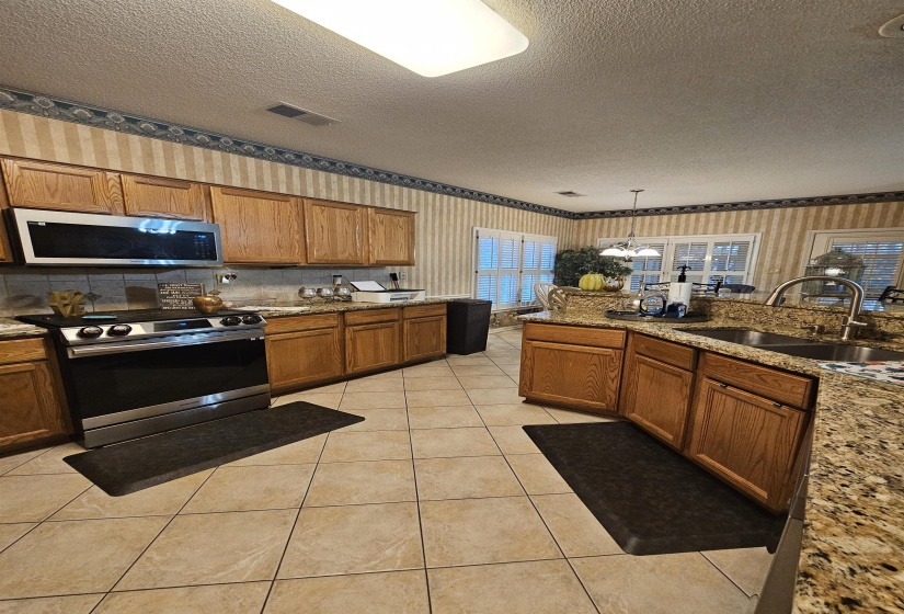 Kitchen with brown cabinetry, a textured ceiling, wallpapered walls, stainless steel appliances, and light stone countertops