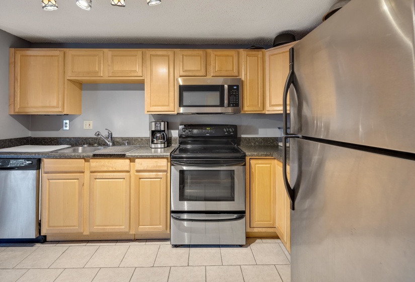 Kitchen featuring stainless steel appliances, dark countertops, light brown cabinetry, and light tile patterned floors
