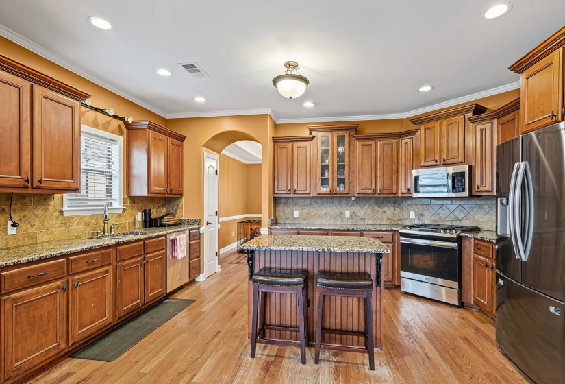 Kitchen with arched walkways, brown cabinetry, stainless steel appliances, light stone counters, and a breakfast bar area