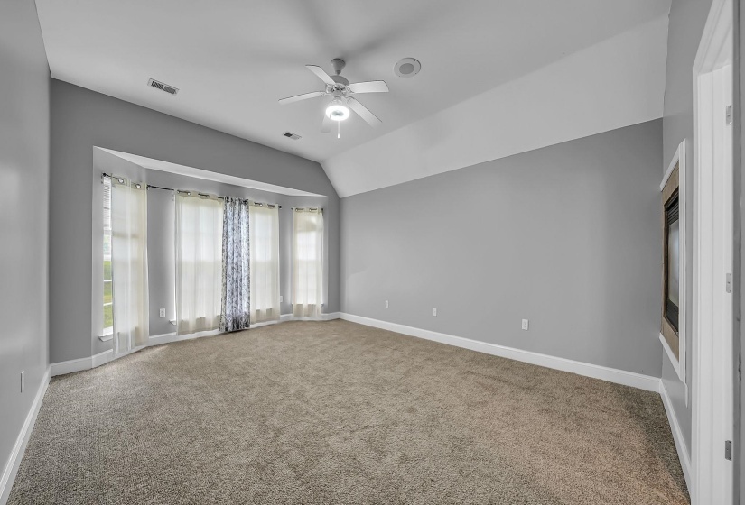 Spacious room featuring vaulted ceilings, a large bay window, ceiling fan with integrated lighting, gray wall paint, and plush carpeting