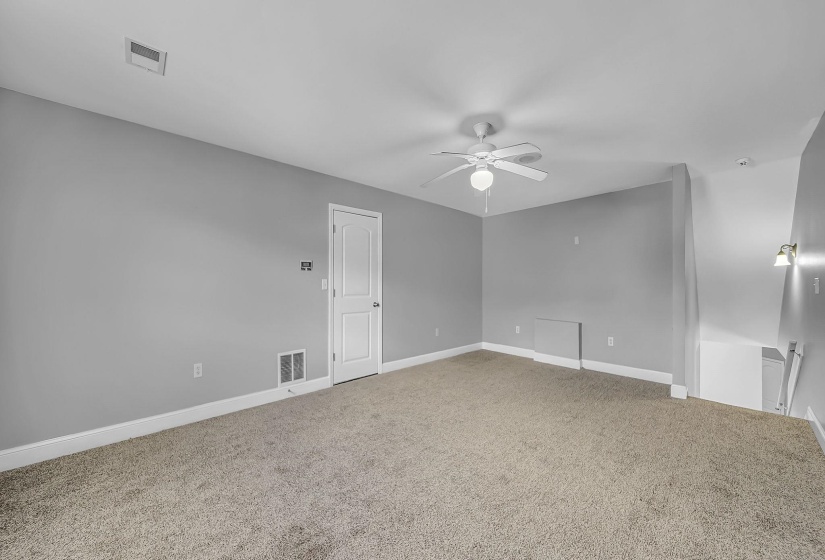 Spacious room with light gray walls, neutral-toned carpet, a ceiling fan with integrated lighting, a white paneled door, and a wall sconce