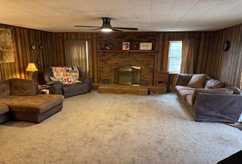 Carpeted living room featuring wooden walls, a ceiling fan, and a brick fireplace