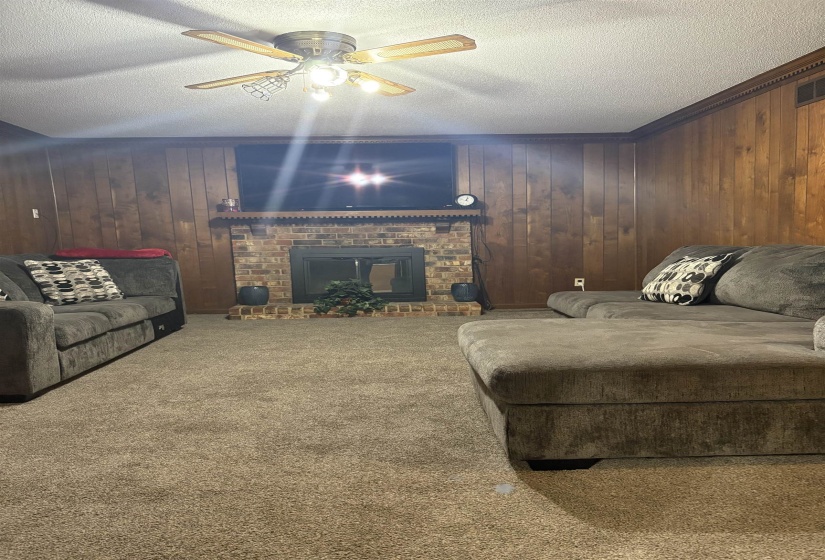 Carpeted living room featuring a textured ceiling, wood walls, a ceiling fan, a fireplace, and ornamental molding