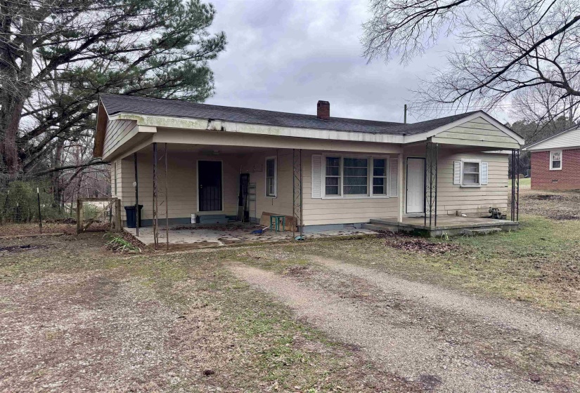 Bungalow with a chimney, an attached carport, dirt driveway, a shingled roof, and a patio