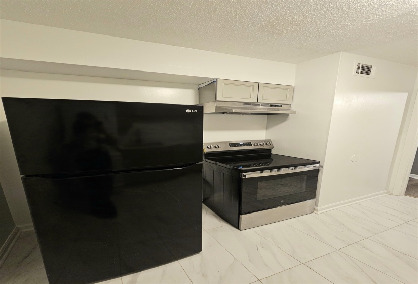 Kitchen with freestanding refrigerator, stainless steel electric stove, a textured ceiling, under cabinet range hood, and light marble finish floors