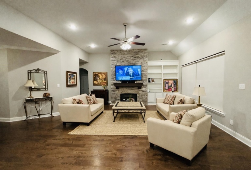 Living room with built in shelves, a stone fireplace, dark wood-style floors, a ceiling fan, and vaulted ceiling