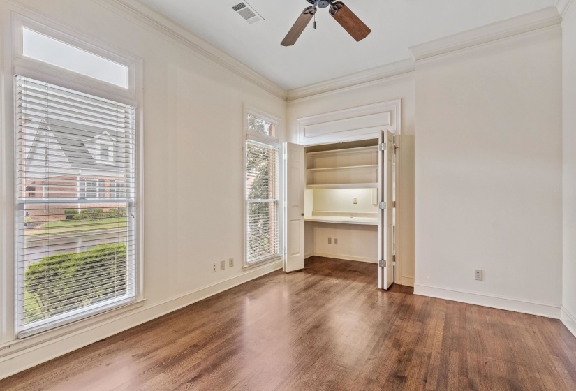 Spacious room featuring wood-finish flooring, two large windows with blinds, crown molding, a ceiling fan, and a built-in desk with shelving and integrated lighting. Could be used as guest bedroom.