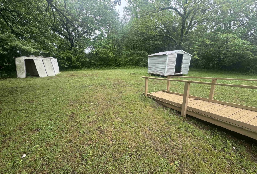 Expansive green space featuring a wooden walkway, two storage sheds, and mature tree-lined borders