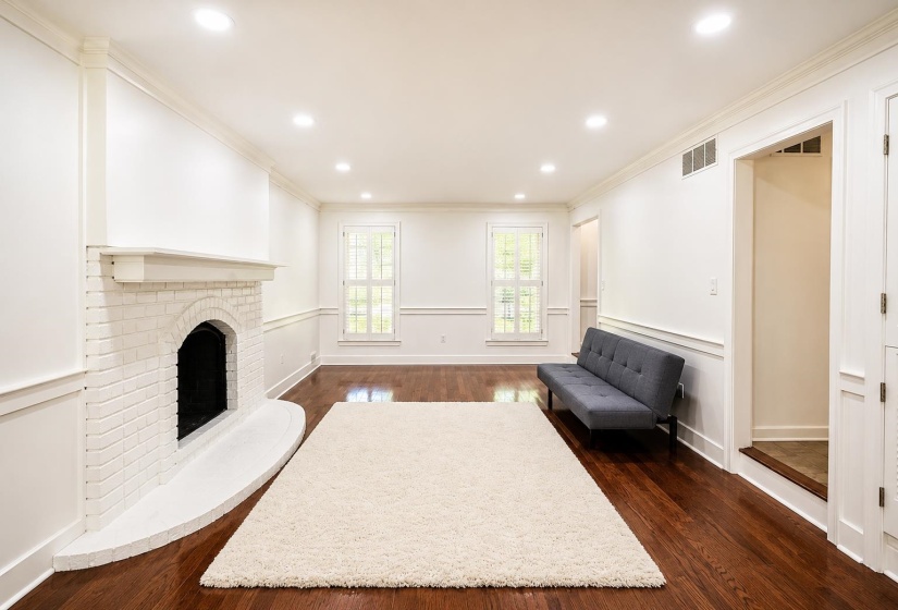 Spacious living area featuring rich hardwood flooring, a white brick fireplace with hearth, recessed lighting, and two windows with plantation shutters
