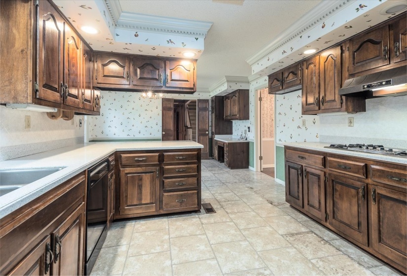 Kitchen with dark brown cabinets, light countertops, crown molding, under cabinet range hood, and wallpapered walls