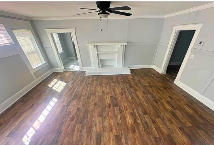 Unfurnished living room with dark wood-type flooring, crown molding, ceiling fan, a fireplace with raised hearth, and a wainscoted wall