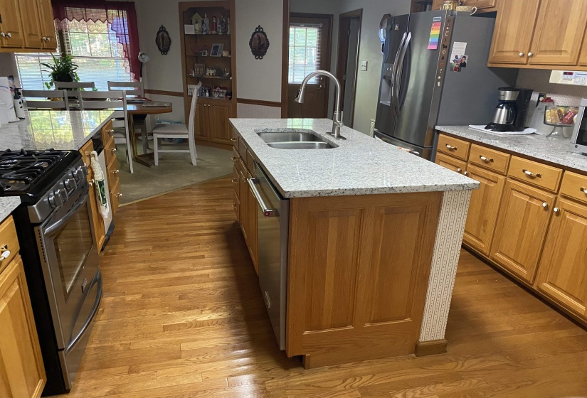 Kitchen featuring brown cabinetry, light granite countertops, appliances with stainless steel finishes, an island with sink, and healthy amount of natural light
