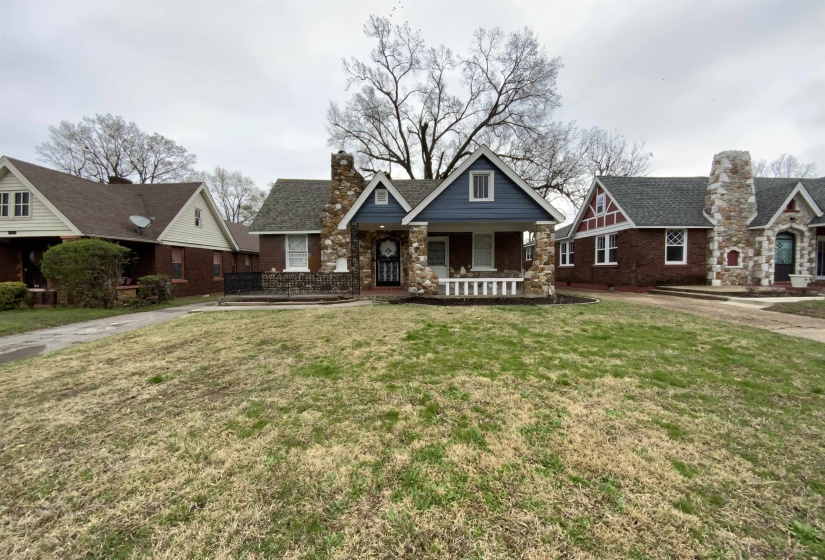 View of front facade with covered porch, a chimney, a front yard, brick siding, and stone siding