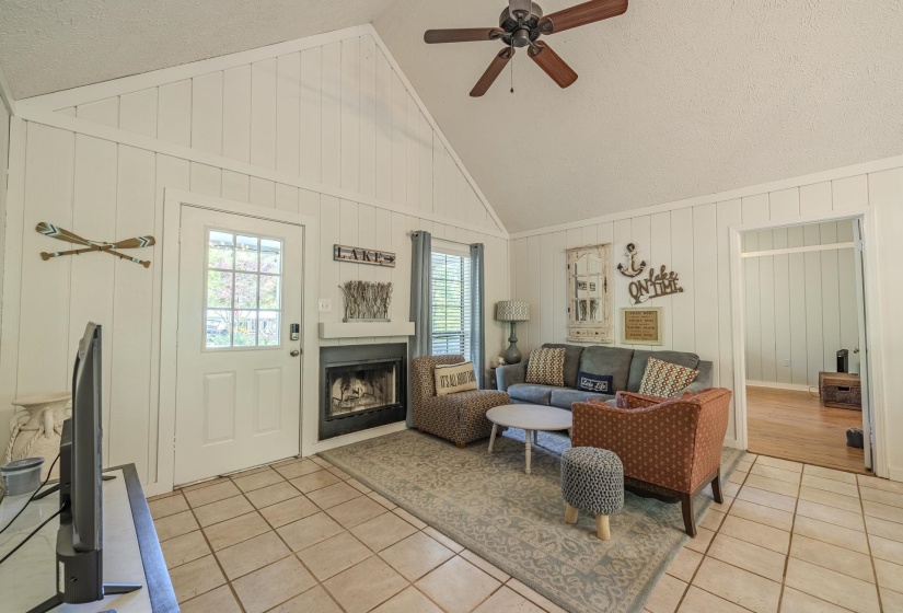 Vaulted ceiling living area featuring tile flooring, a dark-framed fireplace, a white exterior door with glass panes, and a ceiling fan