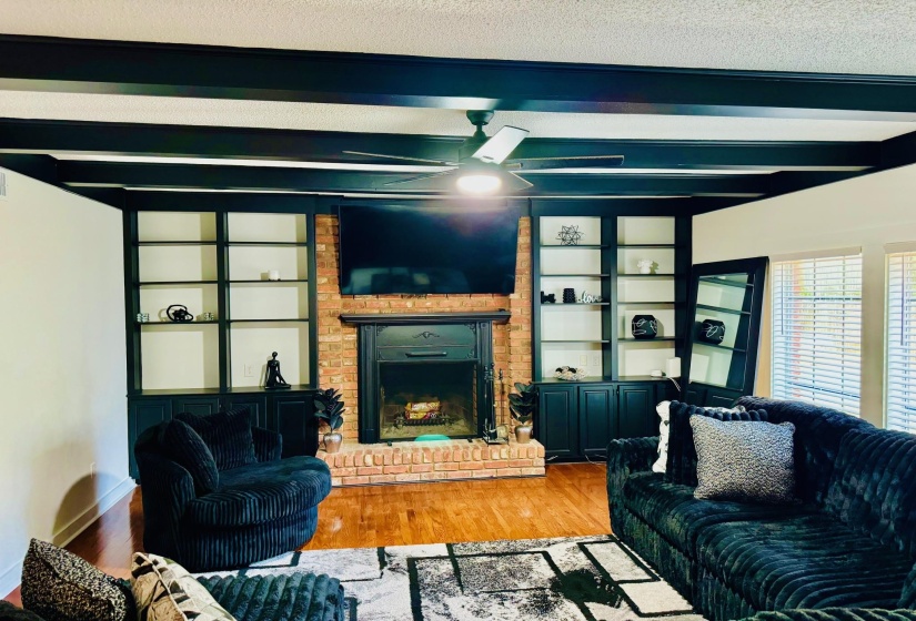 Living area with built in shelves, wood finished floors, a brick fireplace, and beam ceiling