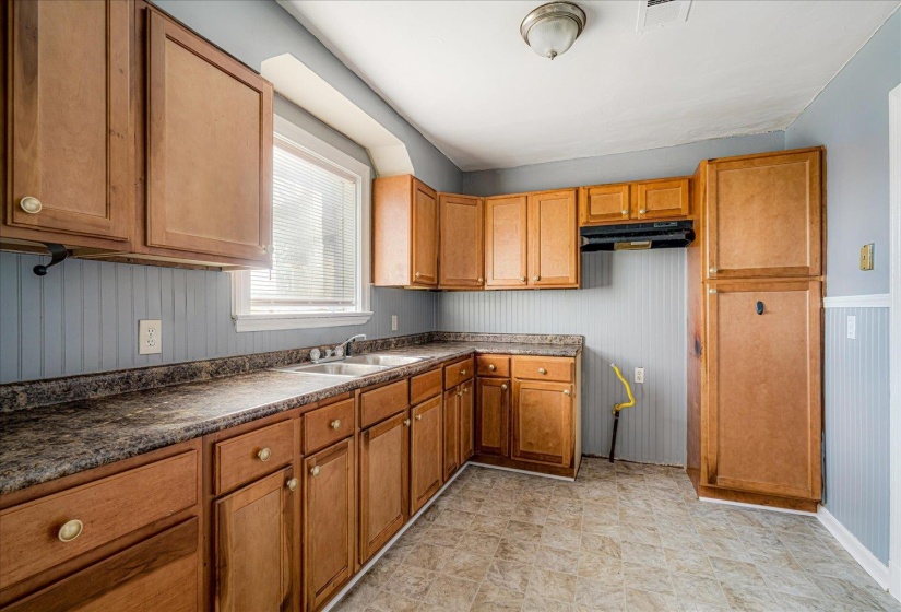 Kitchen with wainscoting, brown cabinetry, dark countertops, under cabinet range hood, and wooden walls