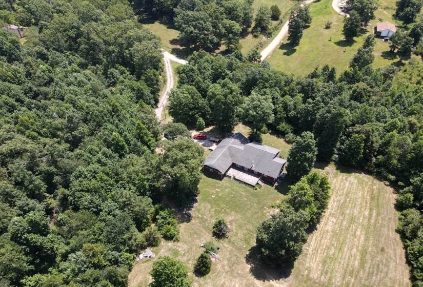 Expansive property featuring a single-story residence with a dark roof, surrounded by mature trees, a partially cleared field, and a winding unpaved drive