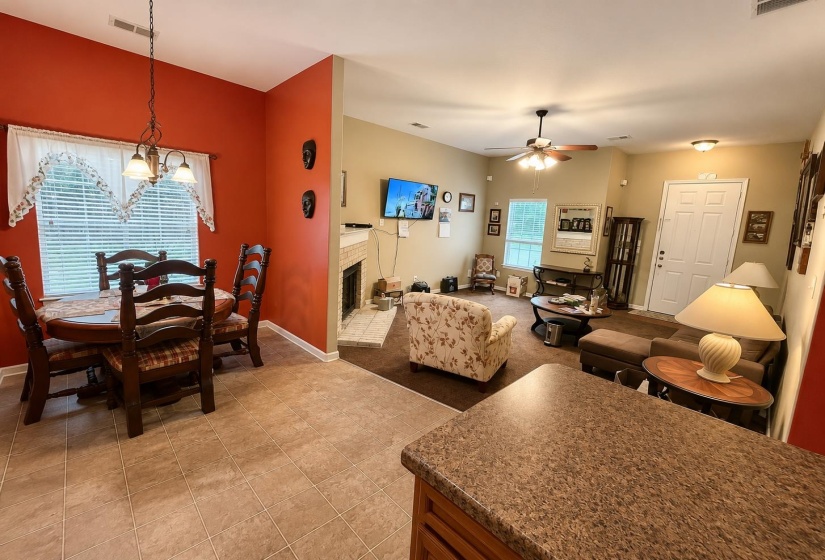 Open-concept living space featuring a dining area with tile flooring and a window, a kitchen counter with a speckled surface, a living room with carpet flooring, and a fireplace with a light-colored surround