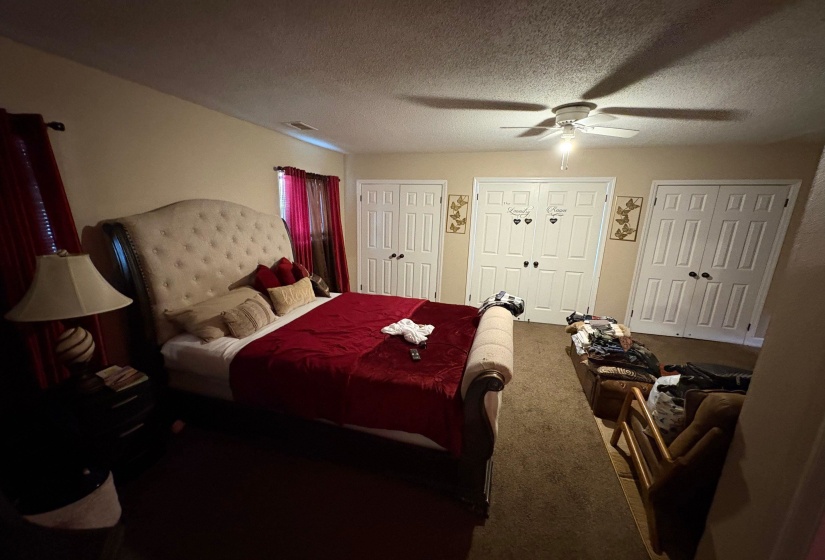 Carpeted bedroom featuring a ceiling fan with light fixture, four white bi-fold closet doors, and two windows with red drapes