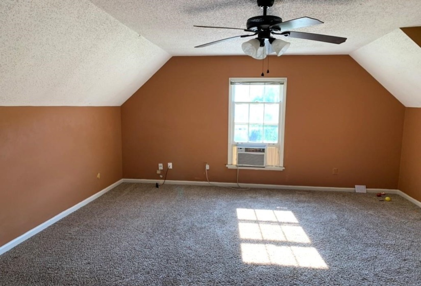 Carpeted room featuring a window with a window-mounted air conditioning unit, a ceiling fan with integrated lighting, and sloped ceilings