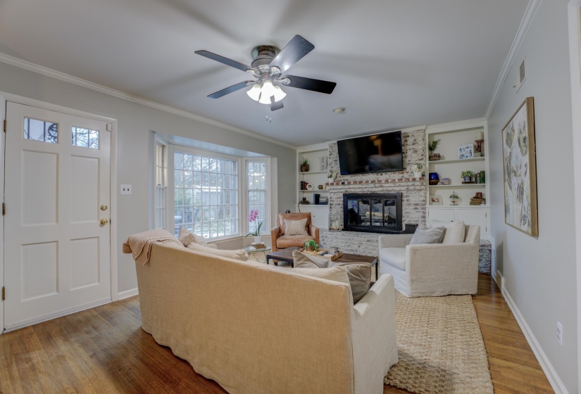 Living area featuring wood-finish flooring, a white paneled entry door with decorative glass, a bay window with multiple panes, a brick-faced fireplace with a mantel, and built-in shelving