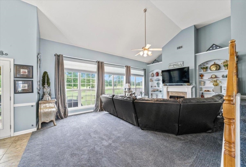 Spacious living room featuring vaulted ceilings, ample natural light from large windows, a fireplace with a mantel, and built-in shelving