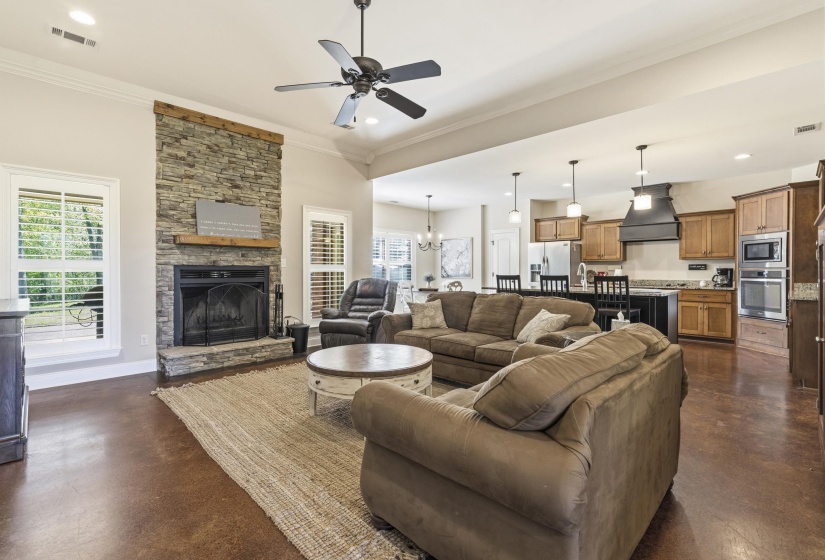 Living area with concrete flooring, a ceiling fan, a stone fireplace, hanging lights, and ornamental molding