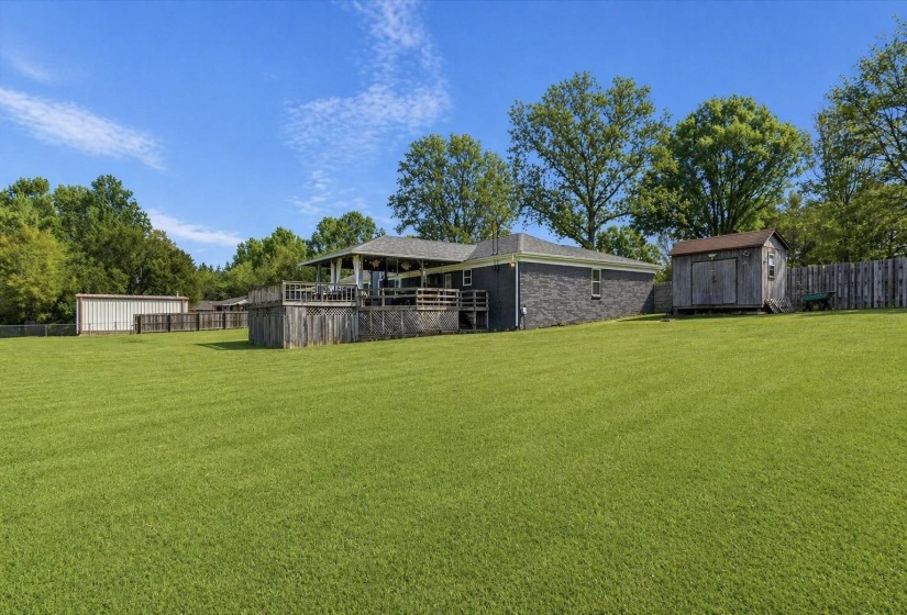 Expansive green lawn surrounds a grey exterior property featuring a covered deck with railings, a wood-sided storage shed, a metal outbuilding, and a wood privacy fence