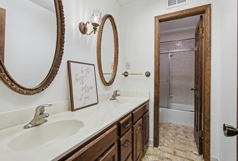 Full bath featuring double vanity, combined bath / shower with glass door, and light stone finish floors