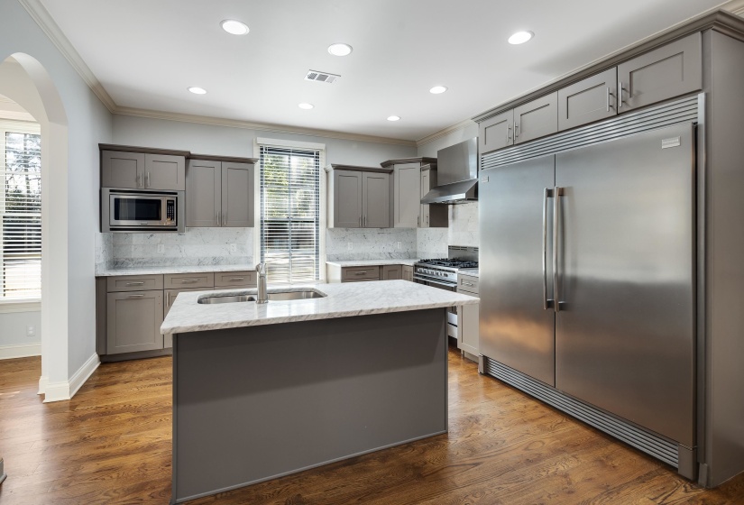 Kitchen with gray cabinetry, built in appliances, dark wood-style flooring, light stone countertops, and a center island with sink