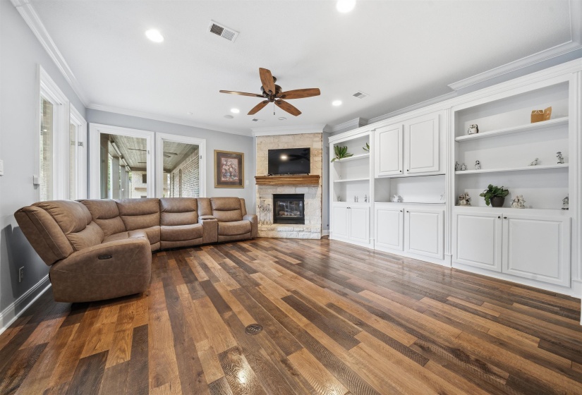 Spacious living area featuring wood-finish flooring, a corner stone-clad fireplace with a wood mantle, custom built-in white cabinetry, recessed lighting, and multiple windows
