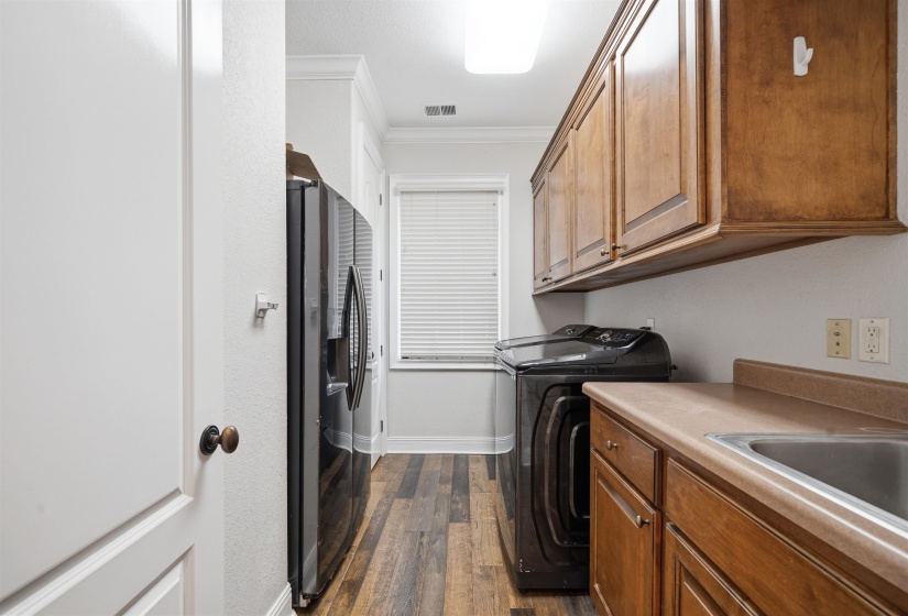 Dedicated laundry area featuring wood-finish flooring, built-in wooden cabinetry, a utility sink, and a window with blinds