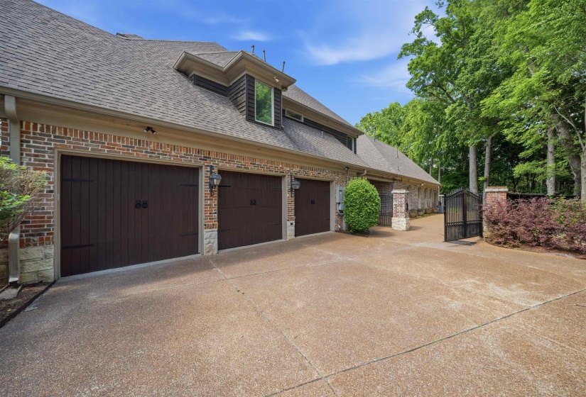 Three dark wood-style garage doors with decorative hardware, brick facade with stone accents, exposed aggregate driveway, dormer window, and a metal entry gate