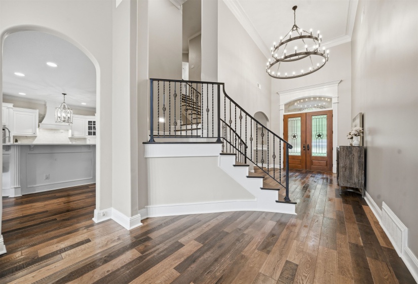 Grand foyer featuring multi-toned wood-finish flooring, a prominent staircase with wrought iron balusters, and a large tiered chandelier