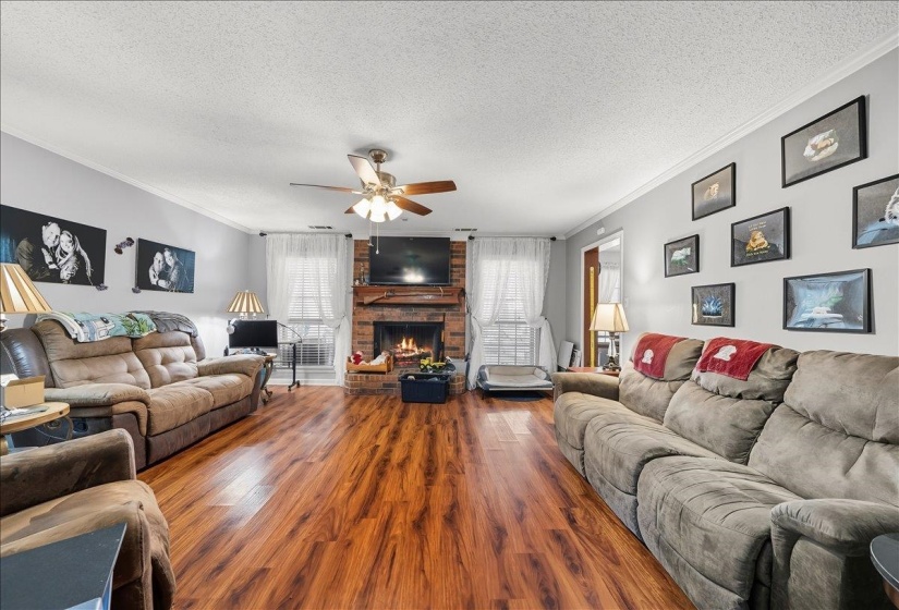 Living area with ornamental molding, wood finished floors, a brick fireplace, a textured ceiling, and ceiling fan